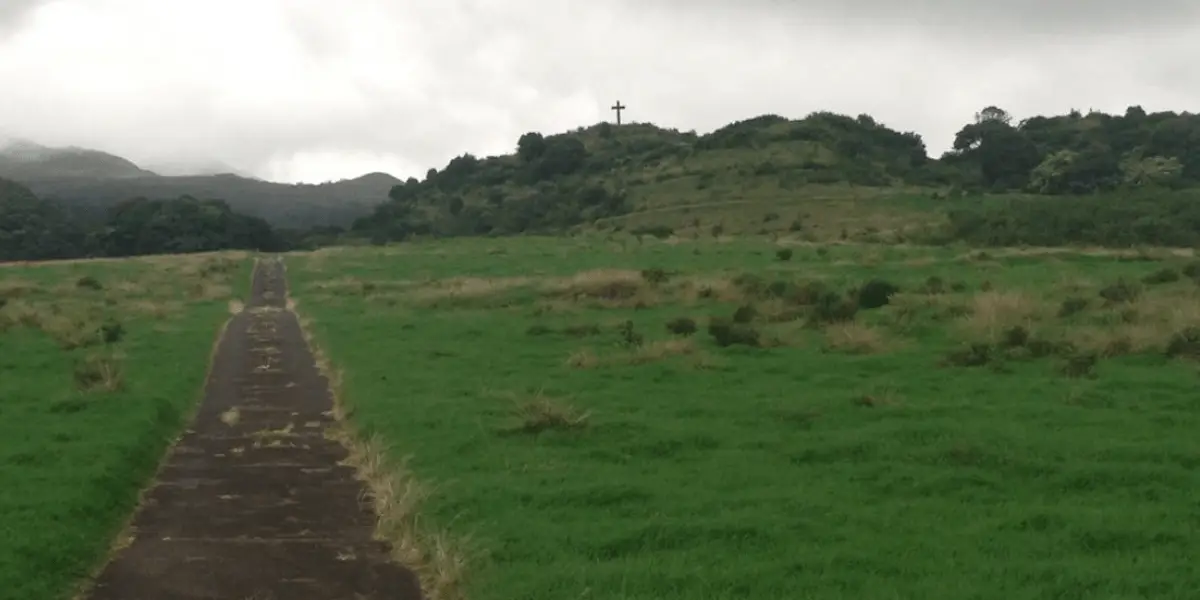 A narrow pathway cuts through a grassy field, leading hikers towards a hill with a cross at its peak. The scene is under a cloudy sky, with dense greenery and trees in the background.