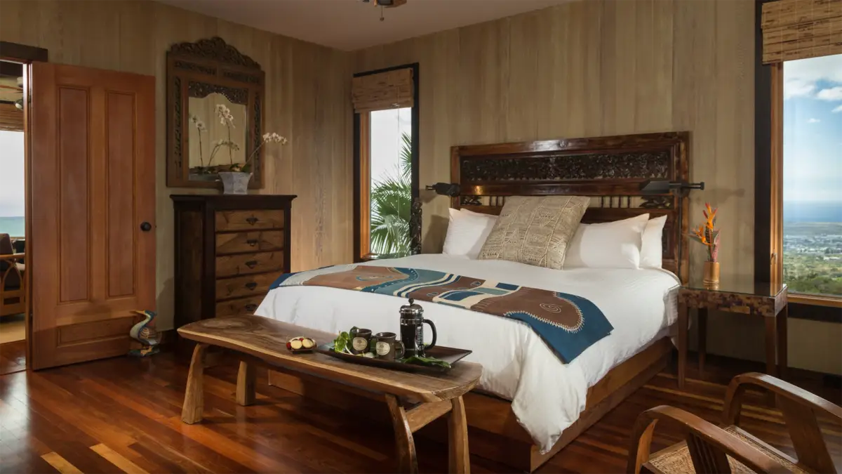 A cozy bedroom with a wooden theme features a large bed adorned with white linens and a patterned blanket, reminiscent of the best big island luxury hotels. A wooden bench sits at the foot of the bed, while a tall dresser with a mirror stands against the wall. The room boasts wooden floors and a view of greenery outside.