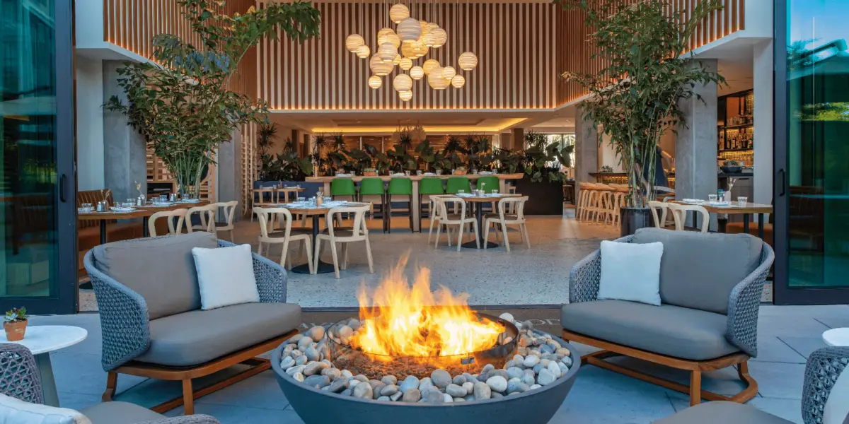 The outdoor seating area of this modern restaurant, one of the best hip hotels in the Bay Area, features a round fire pit with a flame surrounded by stones. Cushioned chairs encircle the fire pit amid lush greenery. Inside, you'll find green bar stools, circular paper lanterns hanging from the ceiling, and wooden decor.