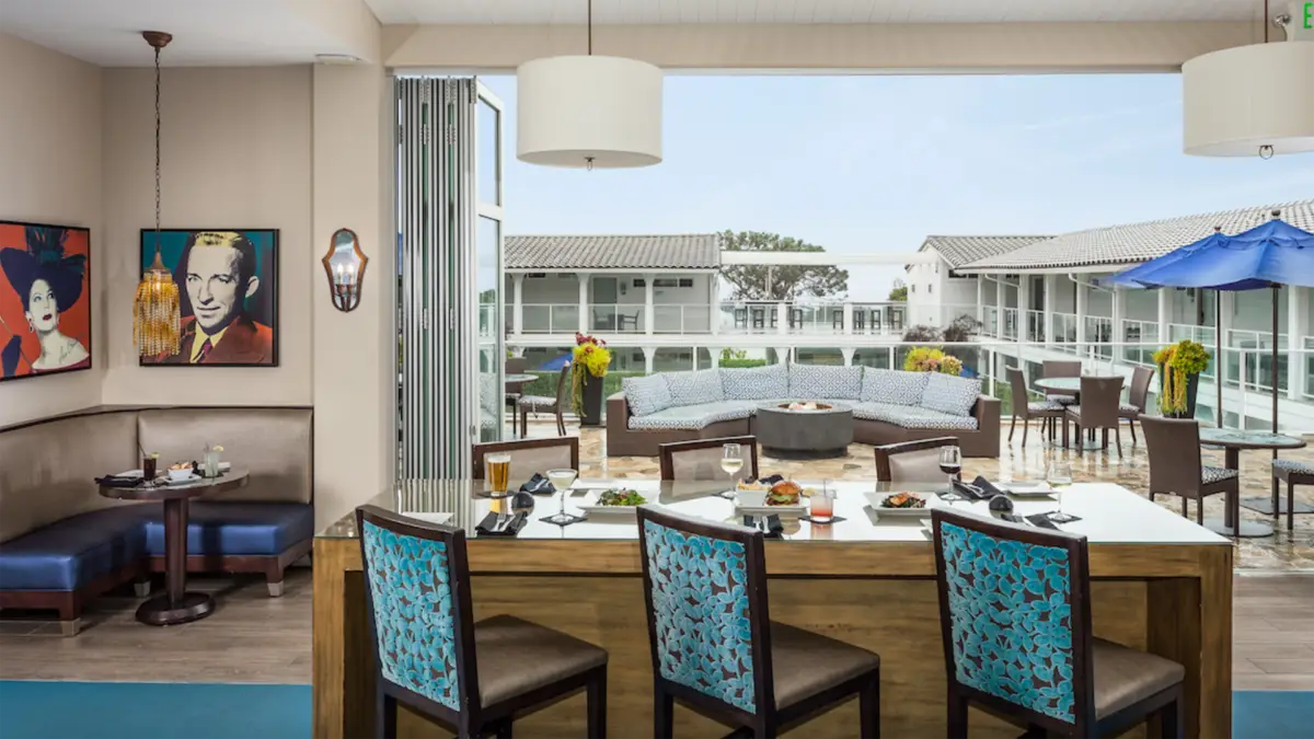 A modern indoor seating area with a kitchen counter and bar stools. A large open window reveals an outdoor patio with cushioned seating and umbrellas. Artwork featuring a woman and a man decorates the wall to the left, reminiscent of some of the best value hotels in San Diego. The setting is bright and inviting.
