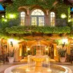 The image depicts a picturesque courtyard restaurant of one of the best group hotels South Bay has to offer, with green ivy-covered walls, warm lighting, and a central stone fountain. Tables are set for dining around the fountain, creating a romantic and inviting atmosphere in the early evening.