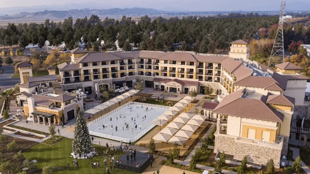 Aerial view of a large resort with buildings surrounding a central outdoor ice skating rink. The rink is encircled by several shaded seating areas, and a decorated Christmas tree stands nearby. Among the best things to do in Wine Country November 2021, this resort offers breathtaking scenes with trees and a distant mountain range.