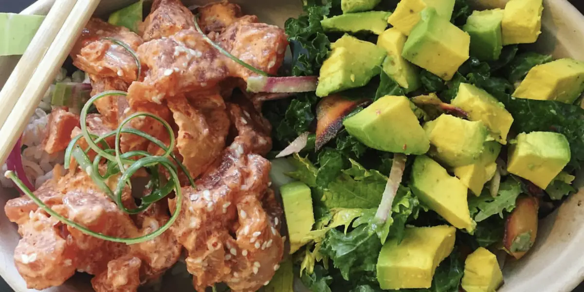 A colorful meal featuring a bowl of lightly sauced salmon poke garnished with green onion strands, sesame seeds, and served with a side of fresh salad made of kale, avocado chunks, and pickled carrots. This dish embodies the flavors you'd find on Maui's best poke stops along the road to Hana. Chopsticks rest on the edge of the bowl.