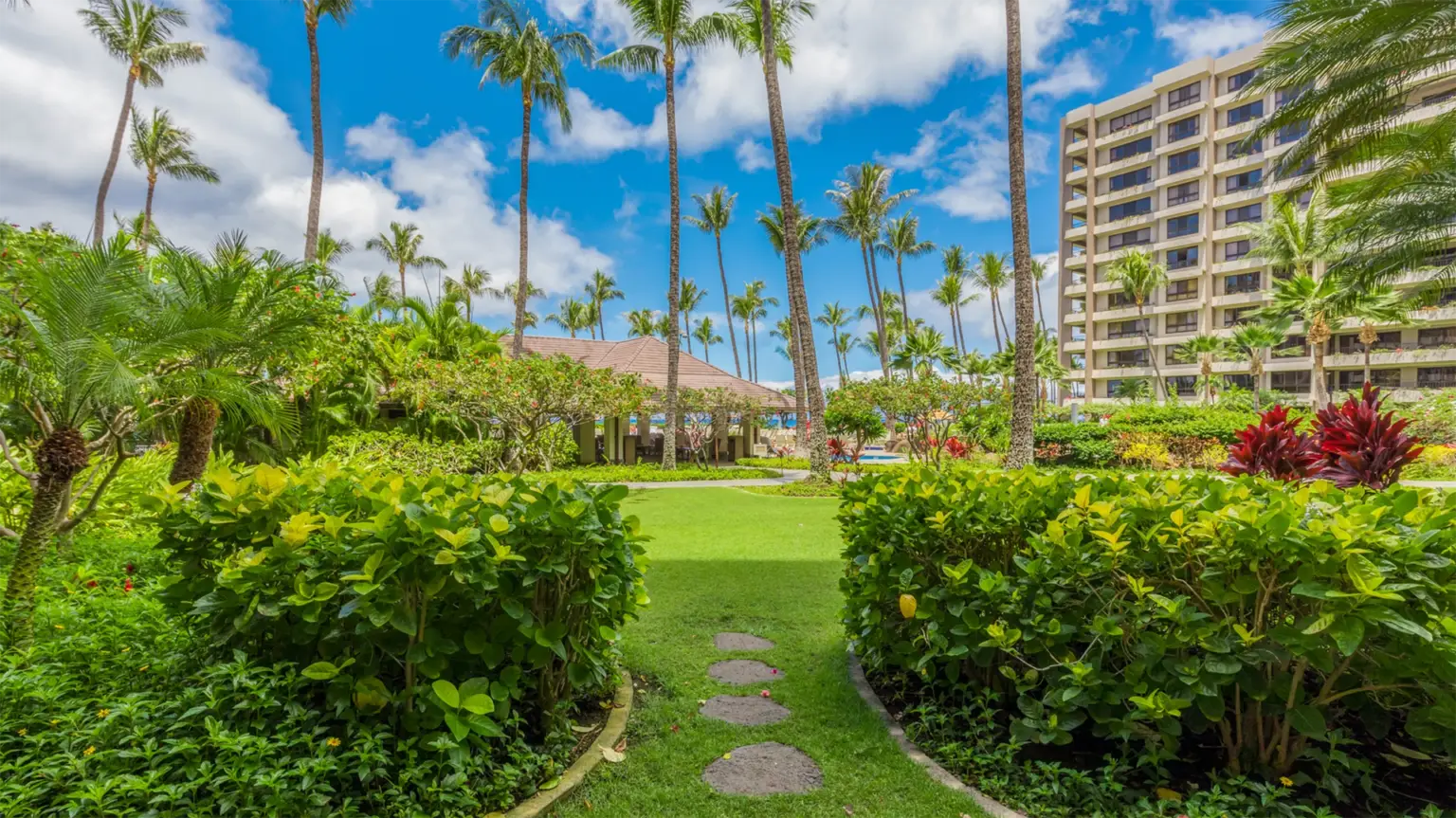 A lush garden with a path of small stepping stones leading toward a building. Tall palm trees and vibrant green foliage surround the area, and a multi-story building is visible on the right side under a bright blue sky with scattered clouds, reminiscent of value hotels Maui visitors adore.