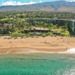 A scenic view of a sandy beach with clear blue waves gently rolling onto the shore. Beachgoers are walking and swimming. Several of the best group hotels Maui offers and lush greenery are visible in the background, with a mountainous landscape rising behind them under a partly cloudy sky.