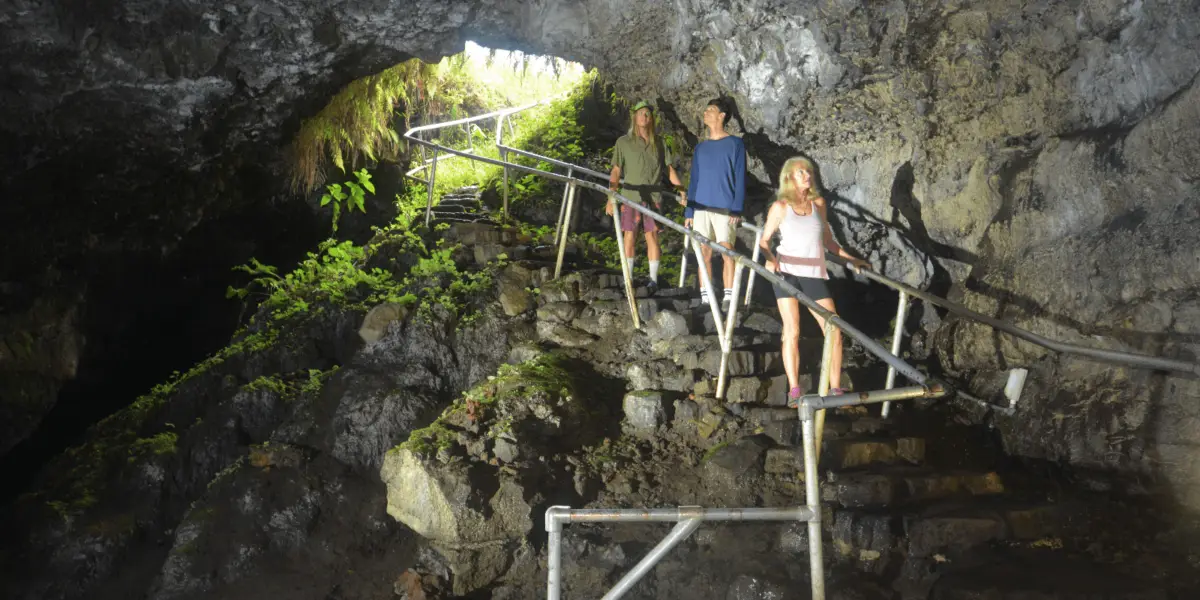 Three people descend a metal staircase inside the rocky Kaeleku Caverns in Hana, with a large opening above. Green plants grow around the entrance, where sunlight filters in, creating a contrast between the brightly lit entrance and the dark interior of this stunning lava tube.