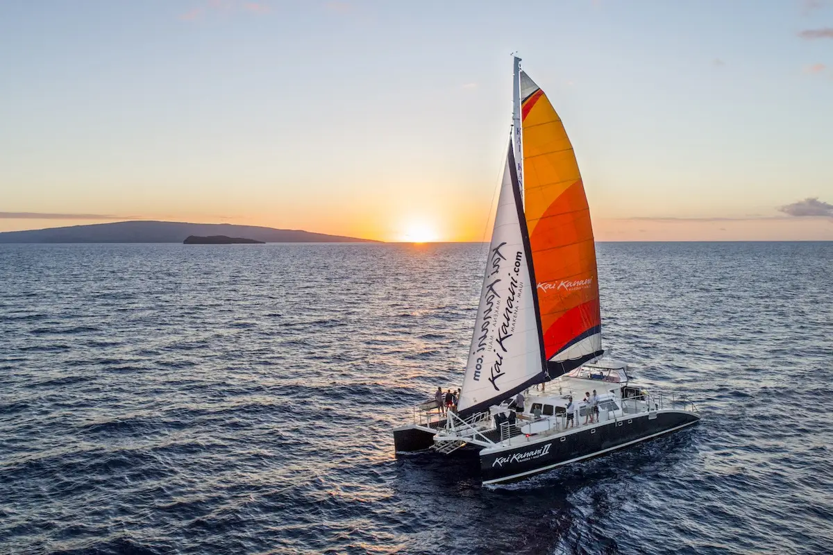 A vibrant sailboat with colorful sails glides on the calm ocean at sunset. The sky is a mix of warm orange and cool blue hues, with a distant island visible on the horizon. Two people are on the boat's deck, enjoying serene surroundings reminiscent of classic Maui activities.