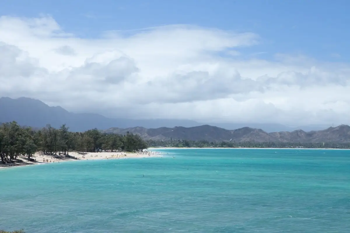 A serene scene at Kailua Beach with turquoise waters gently lapping against a white sandy shore. The beach is bordered by lush green trees and distant mountains under a partly cloudy sky. People are sparsely scattered along the shore, enjoying the tranquil setting.