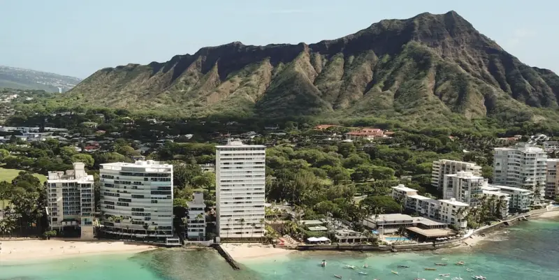 A coastal scene featuring a tranquil Kaimana Beach with turquoise waters in the foreground. High-rise buildings and lush greenery are scattered along the coastline. In the background, a large, rugged volcanic ridge rises against a clear blue sky.