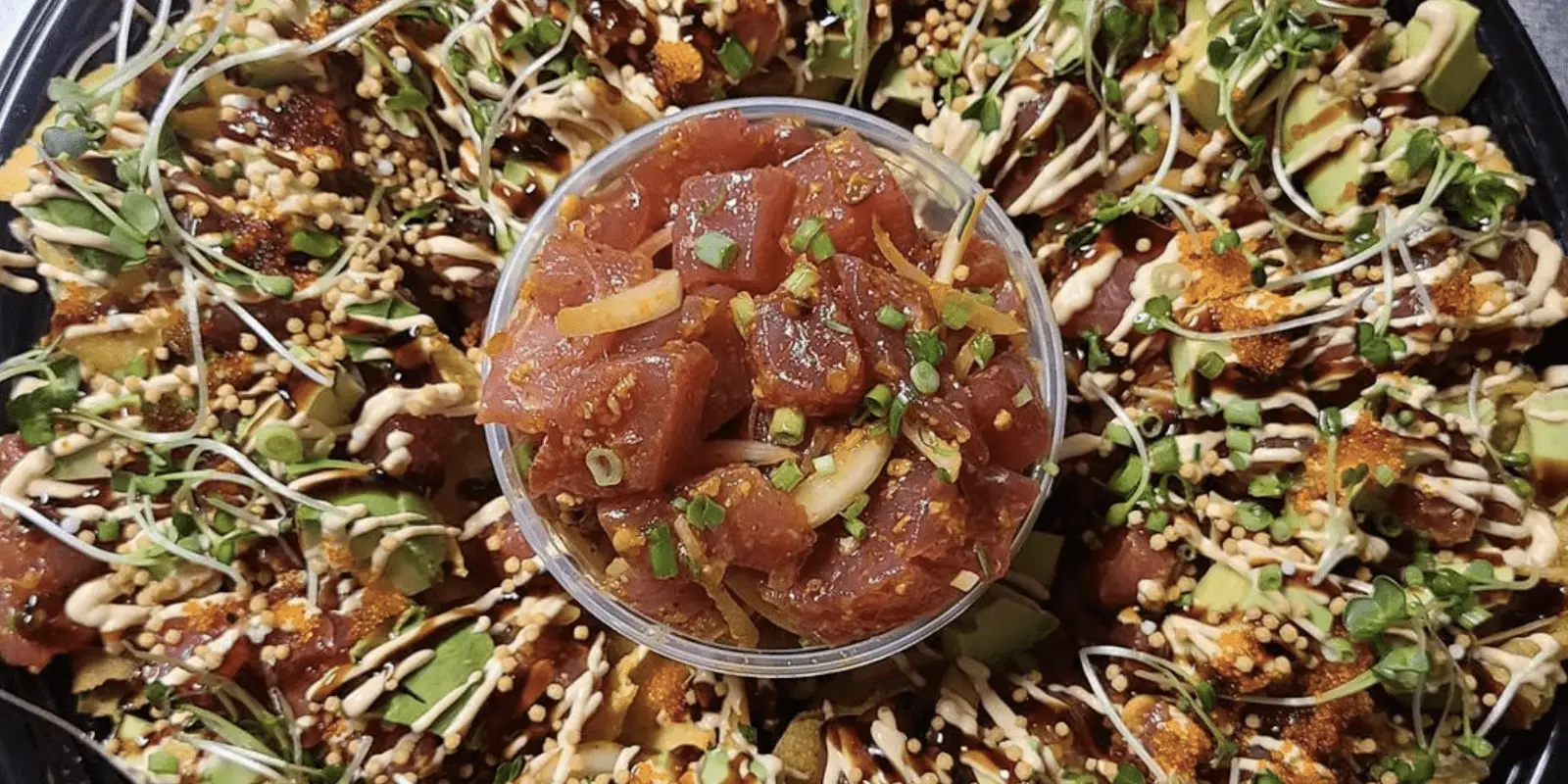 A plate of poke nachos features a center bowl filled with cubed raw fish, sesame seeds, and chopped onions. Surrounding the bowl are tortilla chips topped with avocado, sprouts, and a drizzle of creamy sauce, garnished with spices and green onions—reminiscent of the best poke in Kauai.