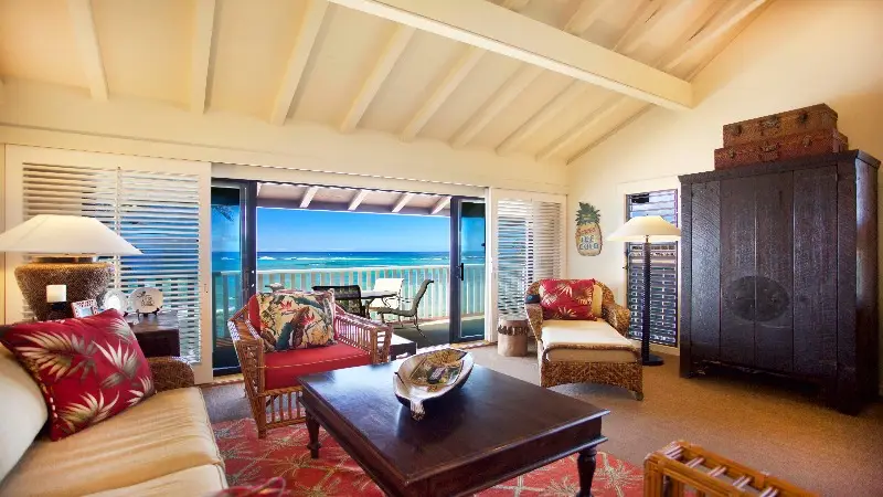 A cozy living room with wooden beams on the ceiling, furnished with a beige sofa, tropical-print cushions, wicker chairs, and a dark wooden coffee table. The room has a large sliding door opening to a balcony with a view of the ocean and a blue sky—reminiscent of the best value hotels Kauai offers.