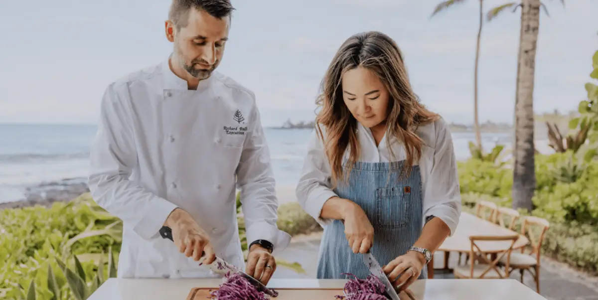 A male chef and a woman are slicing purple cabbage on a cutting board outdoors, enjoying unique culinary spa experiences. The chef in his white coat and the woman in a denim apron are surrounded by lush greenery, with Hawaii Island Resorts' ocean views and palm trees in the background.