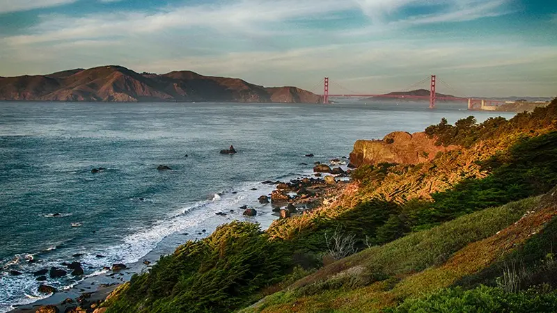 A coastal landscape with the Golden Gate Bridge in the background, partially covered by fog. Rocky cliffs and greenery line the shore, extending into the distance where rugged hills rise under a blue sky with scattered clouds. Waves gently crash against the shoreline—an ideal setting for one of Five Ways to Celebrate July 4th in the Bay Area.