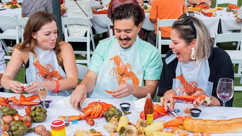 Three people are seated at a table outdoors in Wine Country, enjoying a meal of lobster with various sides like corn and artichokes. They are wearing bibs and laughing together. The table is filled with food, drinks, and condiments, suggesting one of the best things to do in January for a lively and casual dining experience.