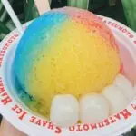 A bowl of colorful shave ice with blue, red, and yellow syrup, garnished with three white mochi balls at the bottom. The bowl is labeled "Matsumoto Shave Ice Hawaii" on its rim. Green leaves are visible in the background, making it clear why it's known as the best shave ice Oahu offers.