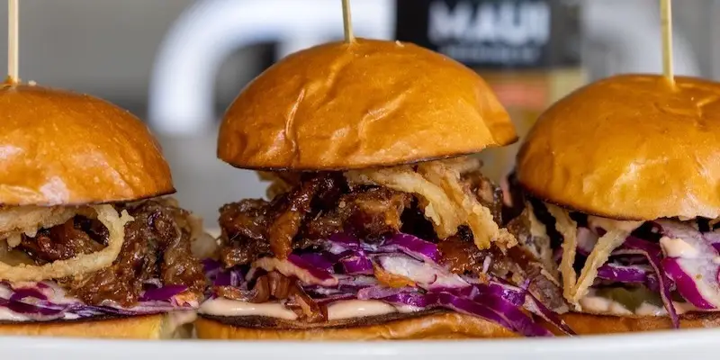 Three gourmet sliders on a plate, each consisting of brioche buns, pulled pork, crispy onion straws, and red cabbage slaw. Skewered with wooden sticks and set against a blurred bottle backdrop, these mouth-watering bites promise to be the highlight of the best happy hour in Waikiki.