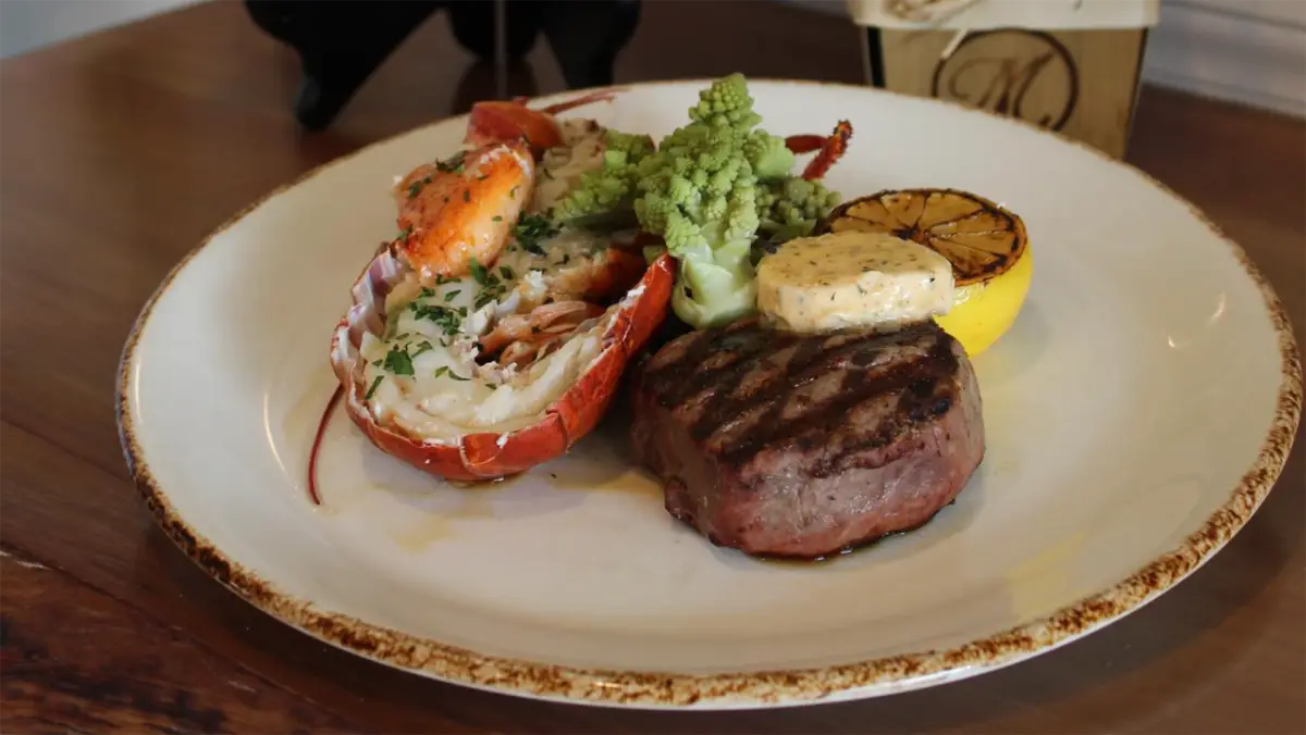 A plate with the best lunch on the Big Island features a gourmet meal: grilled steak topped with herb butter, a lobster tail garnished with herbs, a charred half lemon, and green Romanesco broccoli. The meal is served on a white plate with a brown rim.