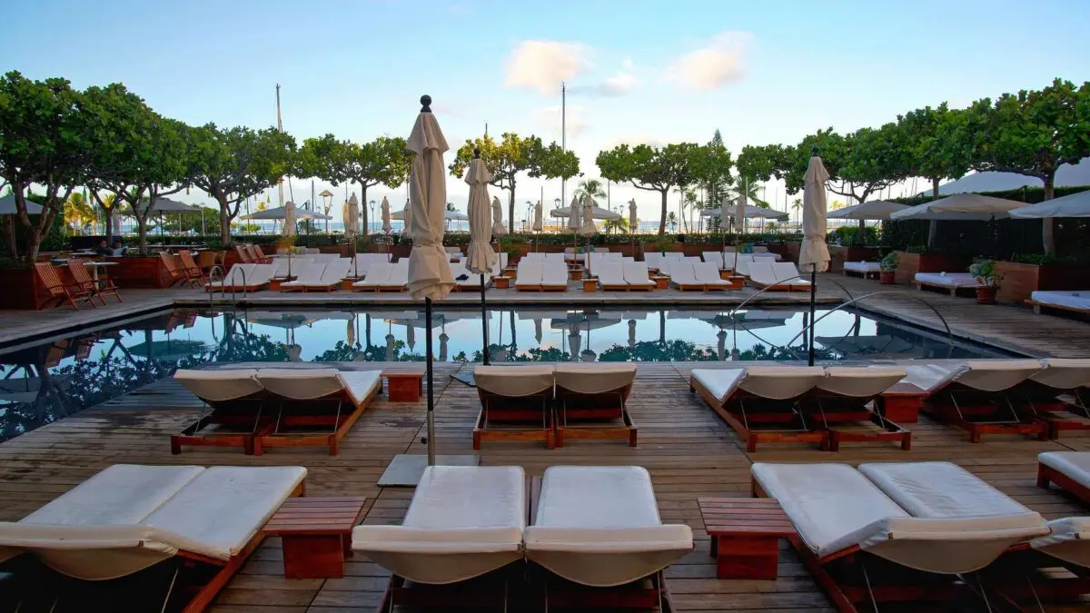 A serene poolside scene featuring several rows of empty, white lounge chairs and umbrellas, surrounding a rectangular pool. Trees line the background with sparse clouds in a blue sky. The atmosphere suggests a tranquil, luxurious setting—perfect for group getaways in Oahu.