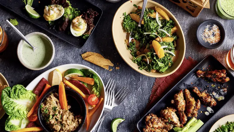 A variety of dishes are spread out on a table at the best happy hour in Wailea, featuring deviled eggs, a salad with orange slices and kale, chicken wings, a small bowl of dip, carrot sticks and lettuce, and a mixed vegetable platter. A fork and a lime wedge are also visible.