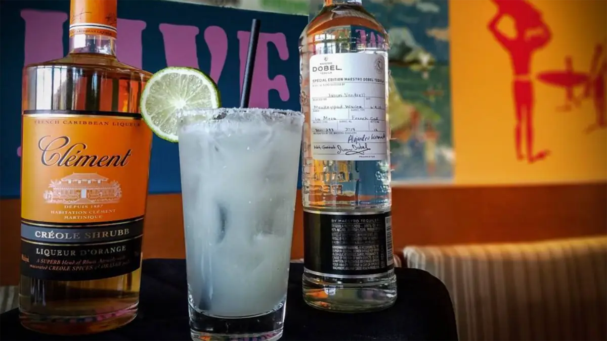 A mixed drink in a glass with a salted rim and a lime slice garnish is centered between two bottles on a table. The bottle on the left is labeled "Clément Créole Shrubb," and the bottle on the right is labeled "Dobel." A colorful sign in the background reads, "Best Margaritas Hawaii.