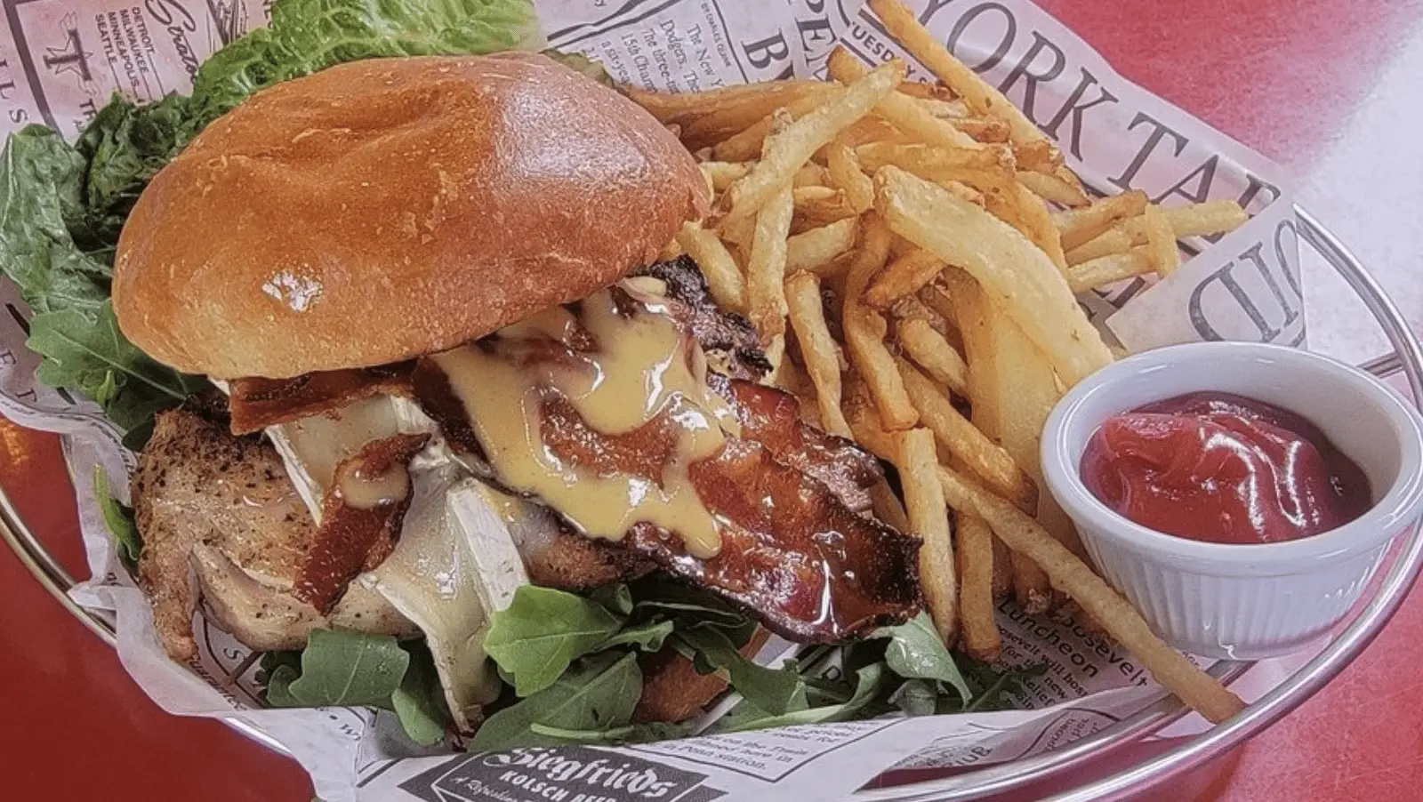 A gourmet chicken sandwich with melted Swiss cheese, crispy bacon, arugula, and honey mustard sauce on a toasted bun, served with a side of golden French fries and a small cup of ketchup, presented on a paper-lined tray—truly the best dinner Tahoe has to offer.