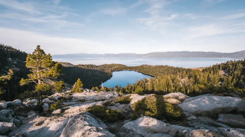 A scenic view of a mountainous landscape with a tranquil lake, one of The 7 Natural Wonders of California, surrounded by dense, evergreen forests. The foreground features rocky terrain and sparse vegetation, while distant mountains are visible under a clear blue sky with scattered clouds.