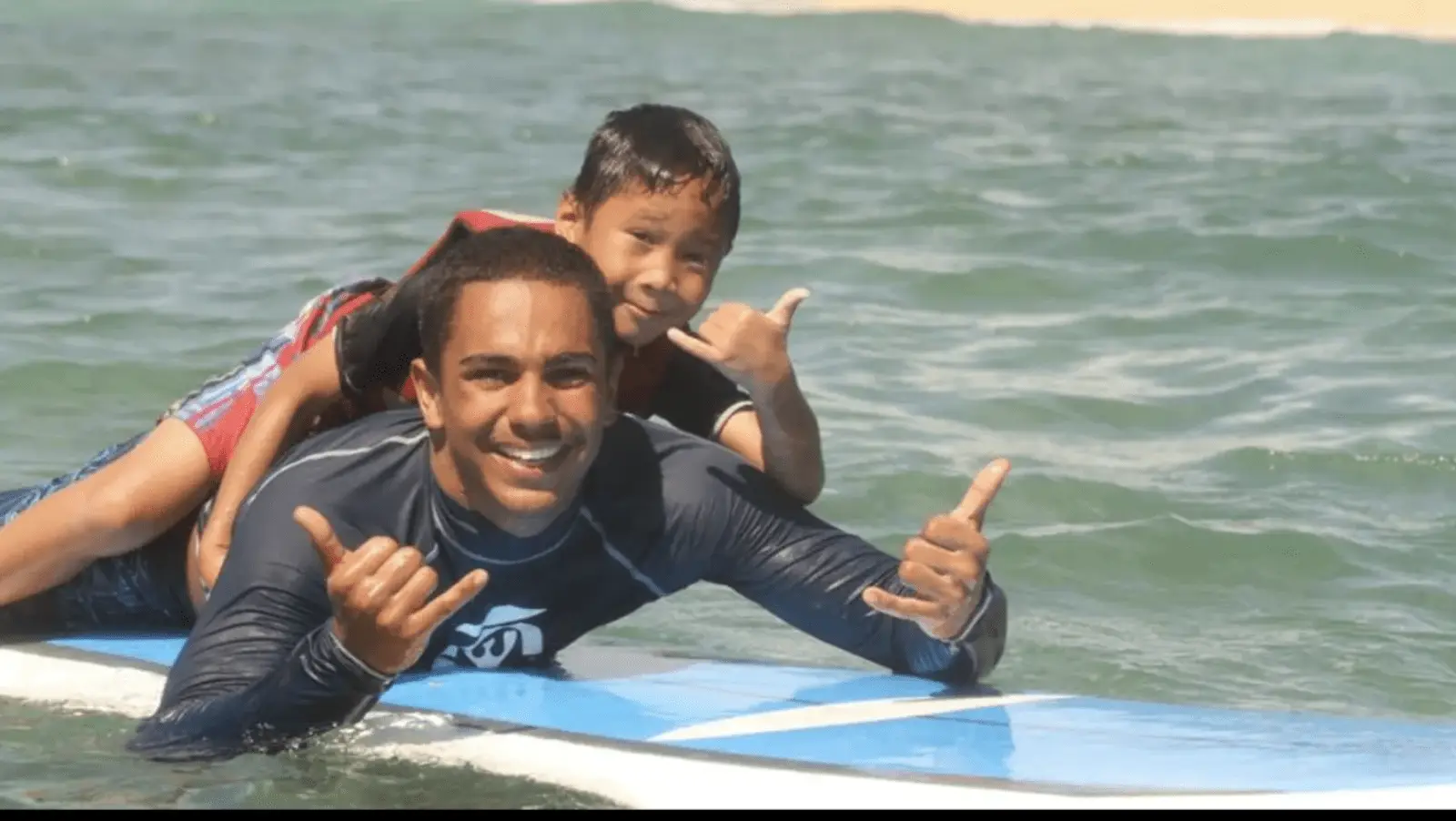 A man and a young boy lie on a surfboard in the ocean. The man in a black rash guard gives a "shaka" hand sign with both hands, smiling widely. The boy, wearing a red life vest, is on the man's back, also making a "shaka" hand gesture with one hand. Just another day at one of the best surf schools on North Shore!
