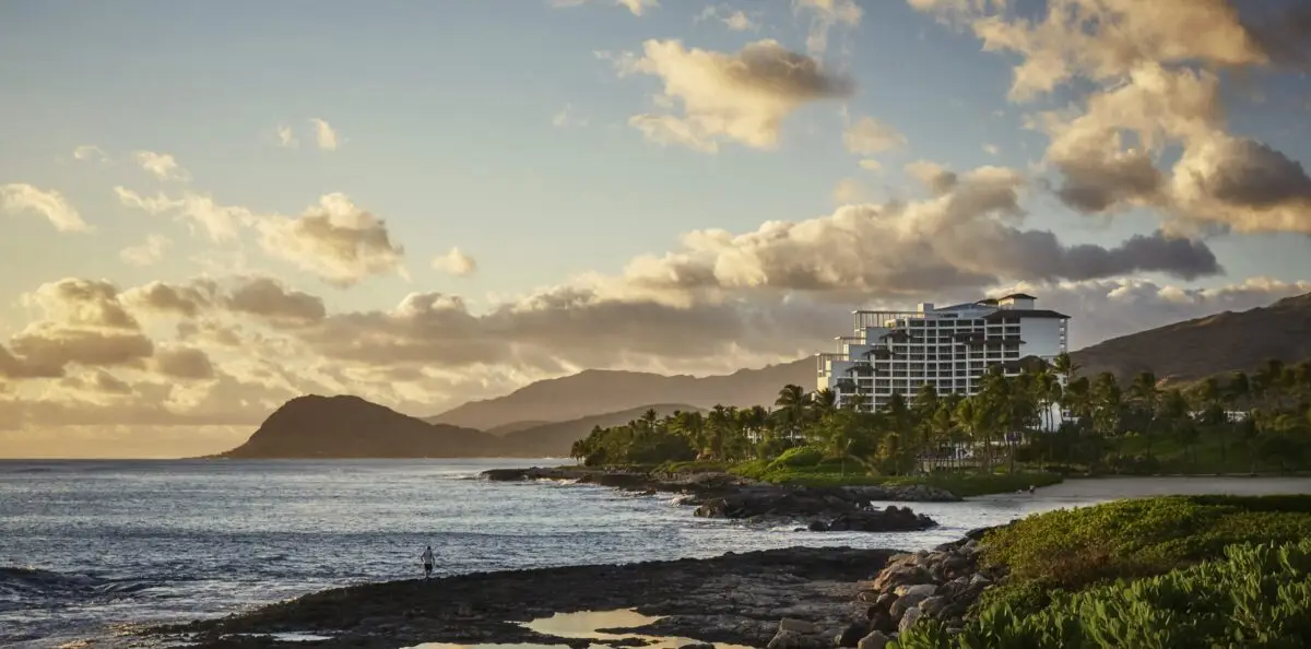 A coastal scene at sunset with a large modern hotel on the right, considered one of the best romantic hotels in Oahu. The hotel is surrounded by lush greenery and palm trees. The ocean has gentle waves, and the mountains in the background are partially shrouded in soft, golden clouds. A person stands near the water's edge.