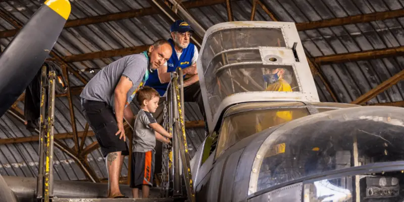 A man and a child stand on a small staircase looking into the cockpit of an old aircraft inside a large hangar, enjoying one of Sonoma County's family-friendly adventures. An elderly man in a blue shirt and cap stands near the cockpit, while another person in a mask observes from behind.