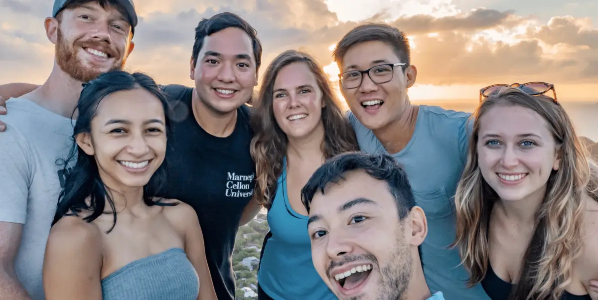 A group of seven people, three women and four men, smile warmly at the camera. They are standing outside with a scenic backdrop of a sunset and cloudy sky. The group appears cheerful and close, capturing one shaka moment together.