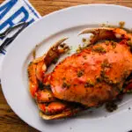 A cooked crab garnished with spices and seasoning is served on a white oval plate with two lemon wedges, showcasing the best seafood in Fisherman's Wharf. The plate rests on a wooden table, accompanied by a napkin with blue text and a crab cracker tool.