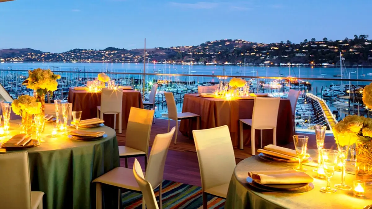 An elegant outdoor restaurant overlooking a marina illuminated by evening lights, perfect for Private Events at Poggio Trattoria. Tables are set with yellow napkins, flower arrangements, and teal tablecloths. White chairs surround the tables while the distant shoreline is dotted with lights from buildings or homes.