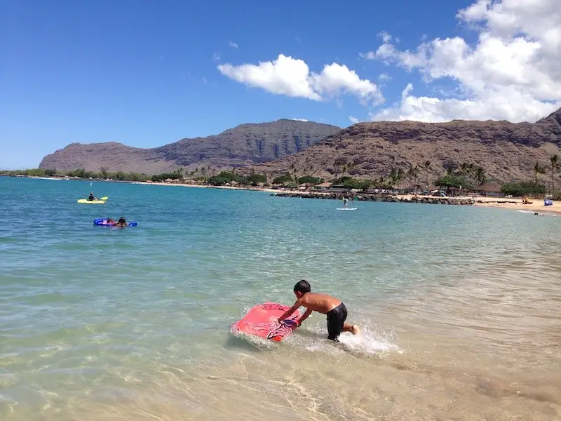 A person pushes a red boogie board into the clear blue water at Pokai Bay's sunny beach. In the distance, other swimmers and beachgoers enjoy the calm sea and surrounding scenic mountains and greenery under a vibrant blue sky with scattered white clouds.
