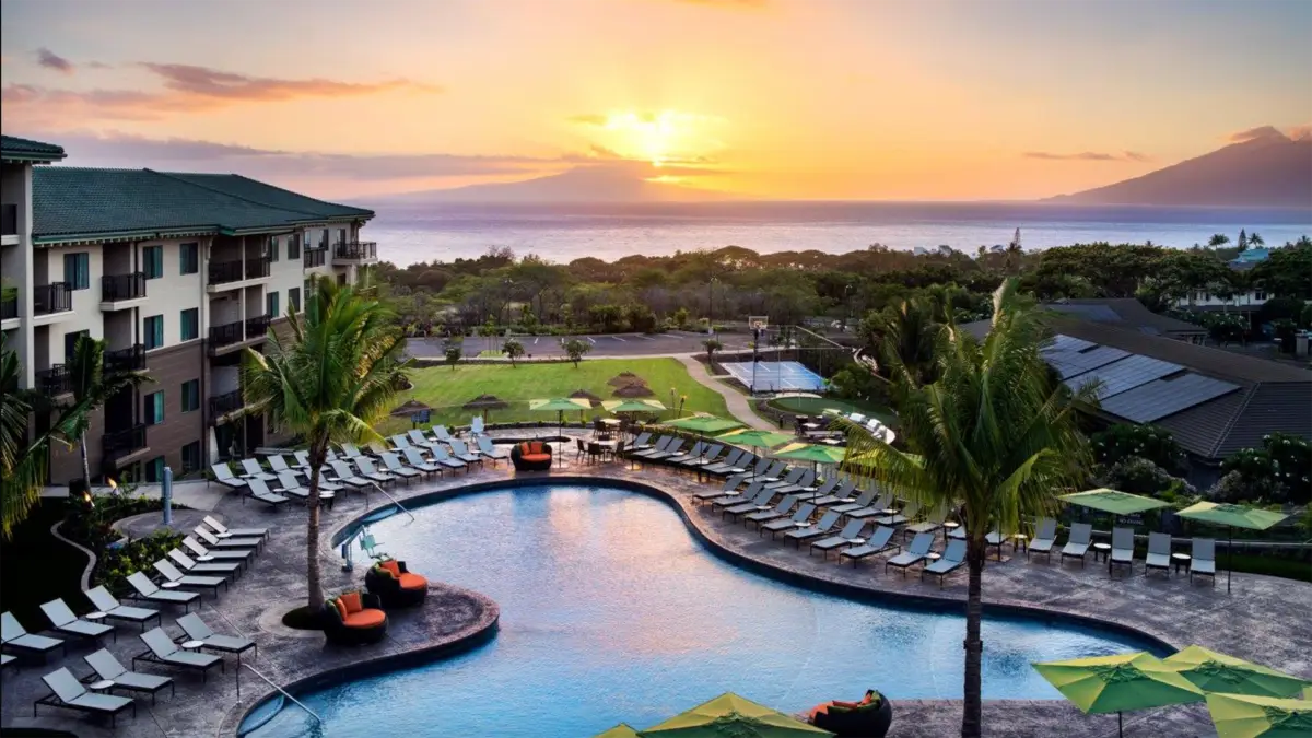 A picturesque sunset view of a tropical resort featuring a large, curved swimming pool surrounded by lounge chairs and palm trees. In the background, there's a vast ocean with an island and majestic mountains under a vibrant, golden sky—reminiscent of the serene beauty found at value hotels in Maui.
