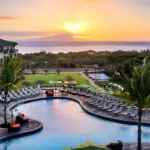 A picturesque sunset view of a tropical resort featuring a large, curved swimming pool surrounded by lounge chairs and palm trees. In the background, there's a vast ocean with an island and majestic mountains under a vibrant, golden sky—reminiscent of the serene beauty found at value hotels in Maui.