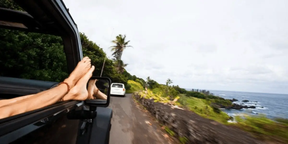 A person with their feet hanging out of a car window is driving along the scenic Road to Hana. Lush green vegetation lines one side of the road, while the ocean with waves crashing against rocks is on the other side. Another vehicle is visible ahead.