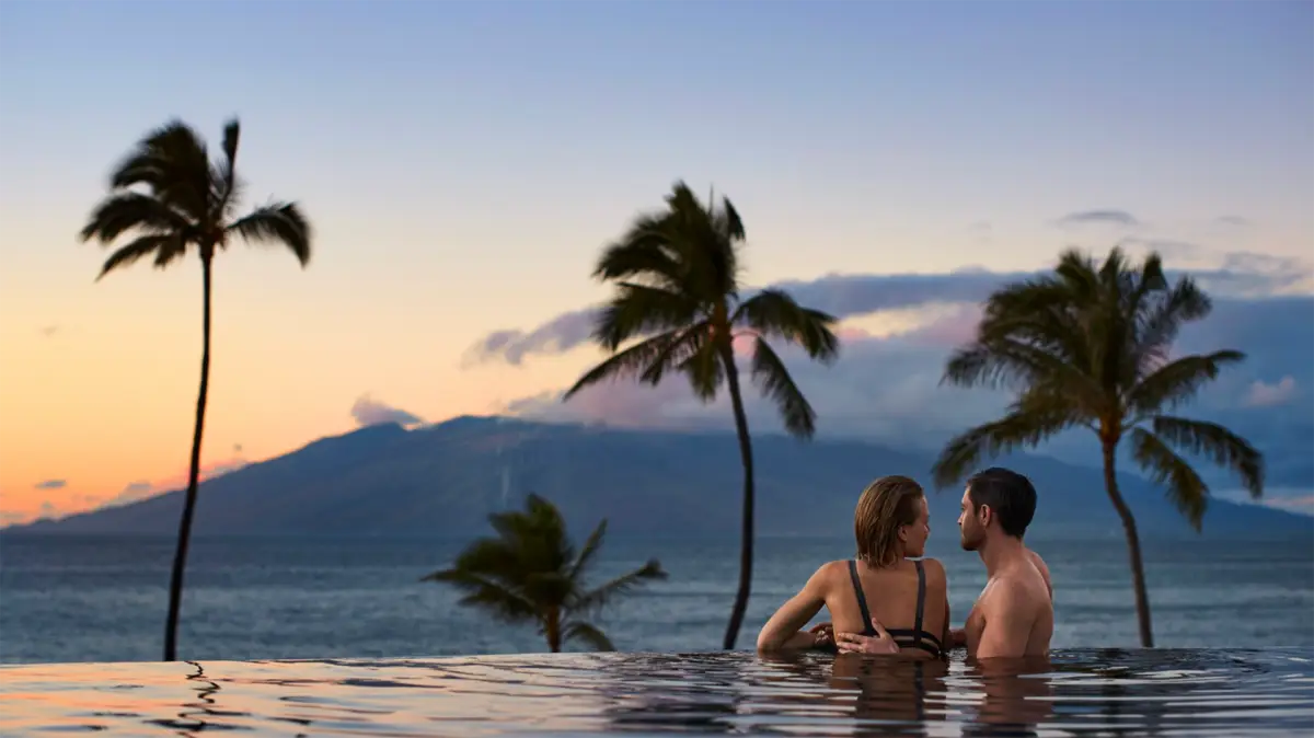 A couple stands in an infinity pool at one of the best romantic hotels in Maui, gazing into each other's eyes. Surrounded by palm trees, they enjoy a scenic view of the ocean and mountains during sunset. The sky is a gradient of blue and orange hues.