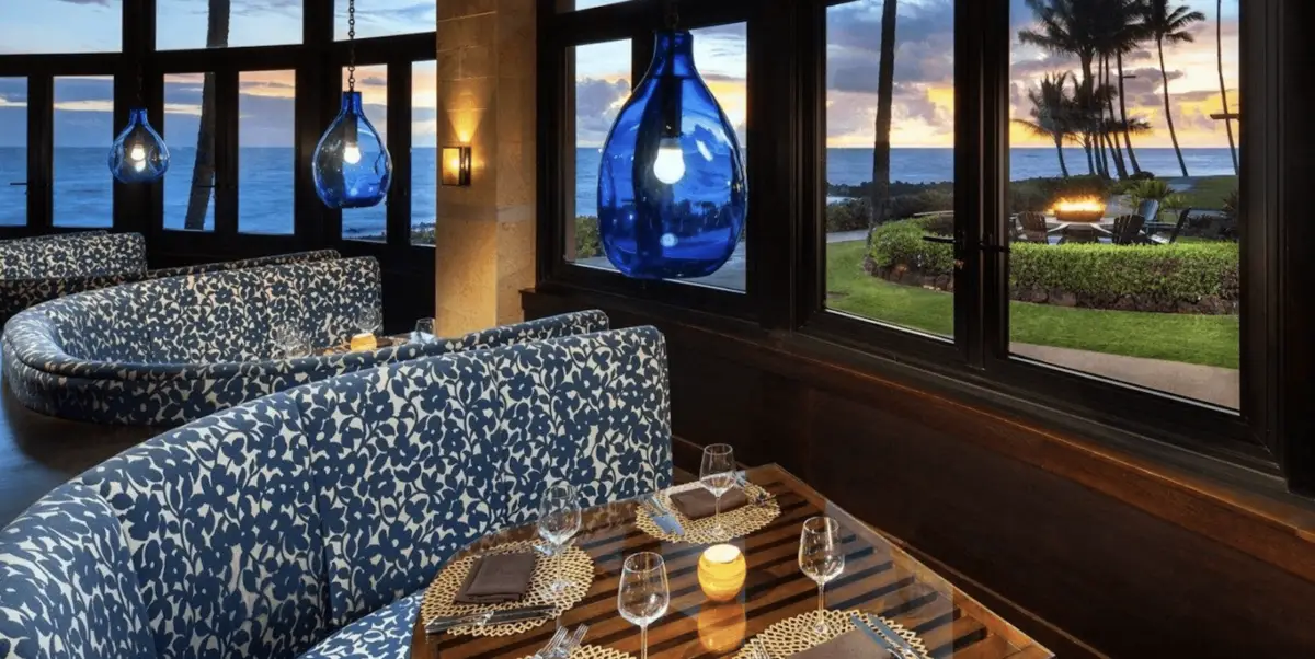 Luxurious restaurant interior with blue patterned seating, glass tables set for a sunset dinner, and large blue pendant lights. Large windows provide a stunning view of a tropical Kauai sunset over the ocean, palm trees, and lush greenery outside.