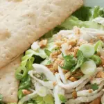 A fresh salad composed of shredded chicken, chopped nuts, and green onions, served on a bed of leafy greens. Two large rectangular crackers are placed on the side in a white bowl, making it one of the best lunch options in North Bay.