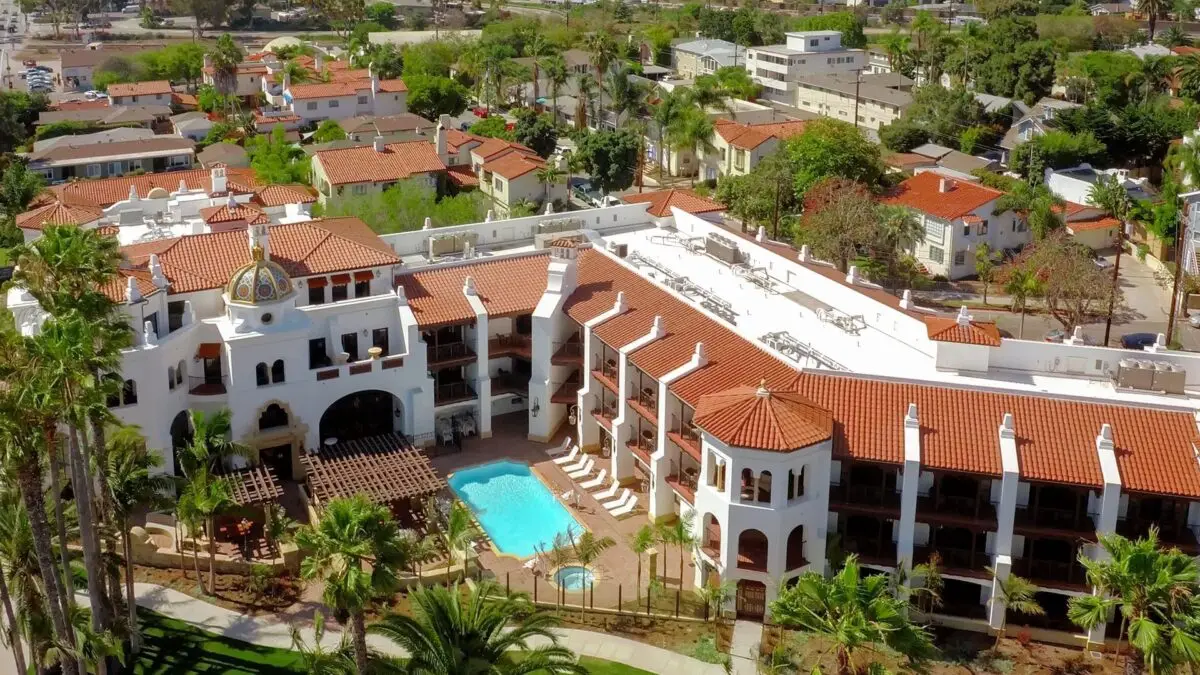 Aerial view of a Spanish-style resort in Santa Barbara featuring white stucco buildings with red-tiled roofs, arched windows, and a central courtyard with a swimming pool. Surrounded by lush green trees and a neighborhood of similar architectural homes, it's one of the best group getaway hotels Santa Barbara offers.