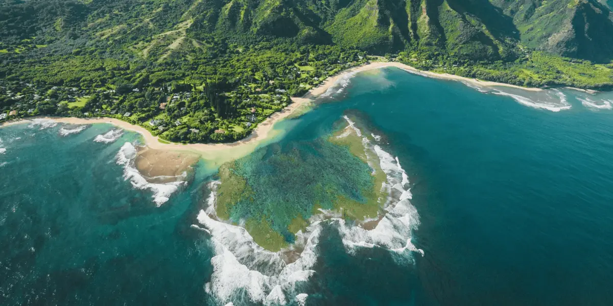 Aerial view of a lush, green coastline with a mountainous backdrop and turquoise waters. A rocky, reef-covered formation is visible in the water near the shore, with waves breaking on the reef. Dense vegetation covers the land, extending to the sandy beach—truly one of the best places to scuba dive on Kauai.