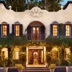 A charming, two-story building with ivy-covered walls and numerous warm-lit windows at dusk, hailed as one of the best romantic hotels in Sonoma. The facade features decorative accents and a crest above the entrance. Surrounded by lush greenery, it exudes a serene, inviting atmosphere.