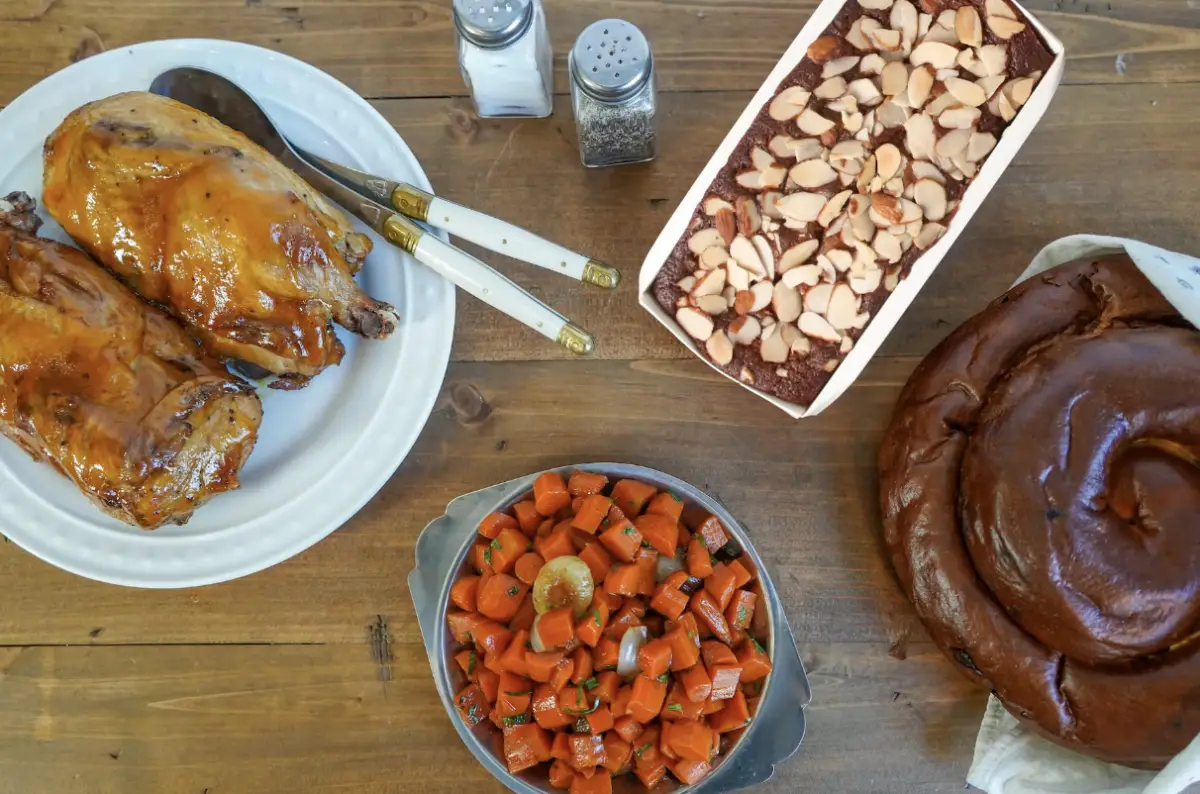 A top-down view of a dining table set for Rosh Hashanah in the Bay Area, featuring roasted chicken on a white plate, baked vegetables in a bowl, a tray of almond-topped cake, and a round loaf of challah bread. Salt and pepper shakers are placed in the background.