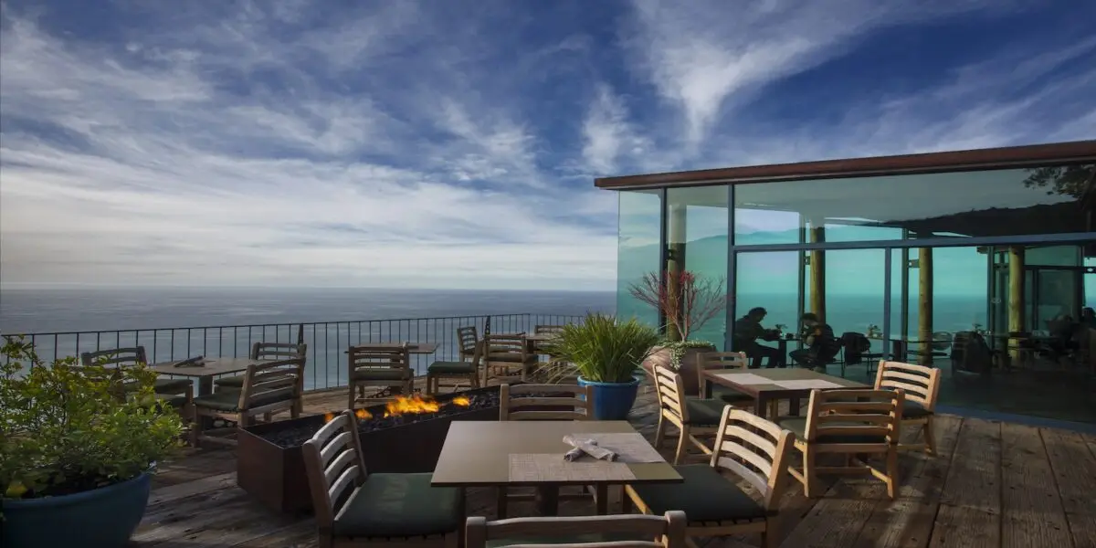 A serene outdoor seating area of one of the best restaurants in Big Sur features tables and chairs, with a modern glass-walled section extending from the main building. A fire pit is surrounded by potted plants, and the open-air deck overlooks a calm ocean under a partly cloudy sky.