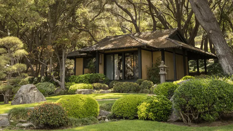 A serene Japanese tea house stands amidst lush greenery and trees, reminiscent of the best spas in Southern California. The traditional structure features sliding shoji doors and a sloping tiled roof. The surrounding garden has neatly trimmed bushes, stone lanterns, and a well-manicured lawn, creating a tranquil setting.