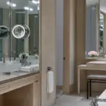 A luxurious bathroom with double sinks, a large mirror with illuminated magnifying mirrors, and fresh flowers. The space features beige tones and modern decor. A vanity area with a small bench and flowers is visible in the background, reminiscent of the best spa hotels San Francisco boasts.