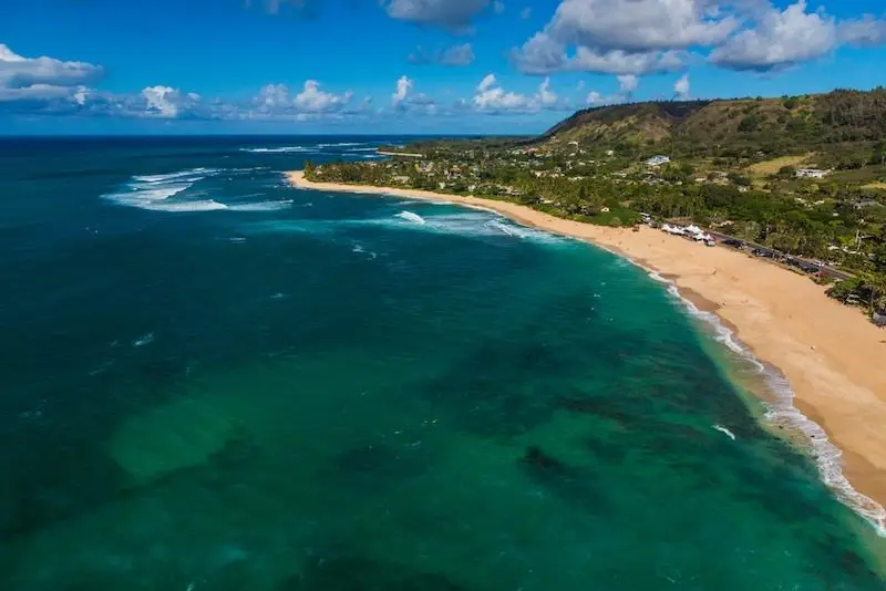 Aerial view of Sunset Beach with golden sand and turquoise waters. The shoreline curves gently, bordered by lush green vegetation and distant hills. Waves gently break along the coast under a partly cloudy blue sky. Some buildings are visible in the distance.