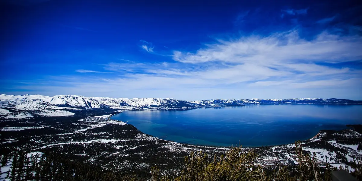 A scenic view of Lake Tahoe, one of the Best Places to Visit, surrounded by snow-capped mountains under a bright blue sky with scattered clouds. The calm, reflective surface of the lake contrasts with the rugged, snowy terrain and dark forested areas on the slopes.