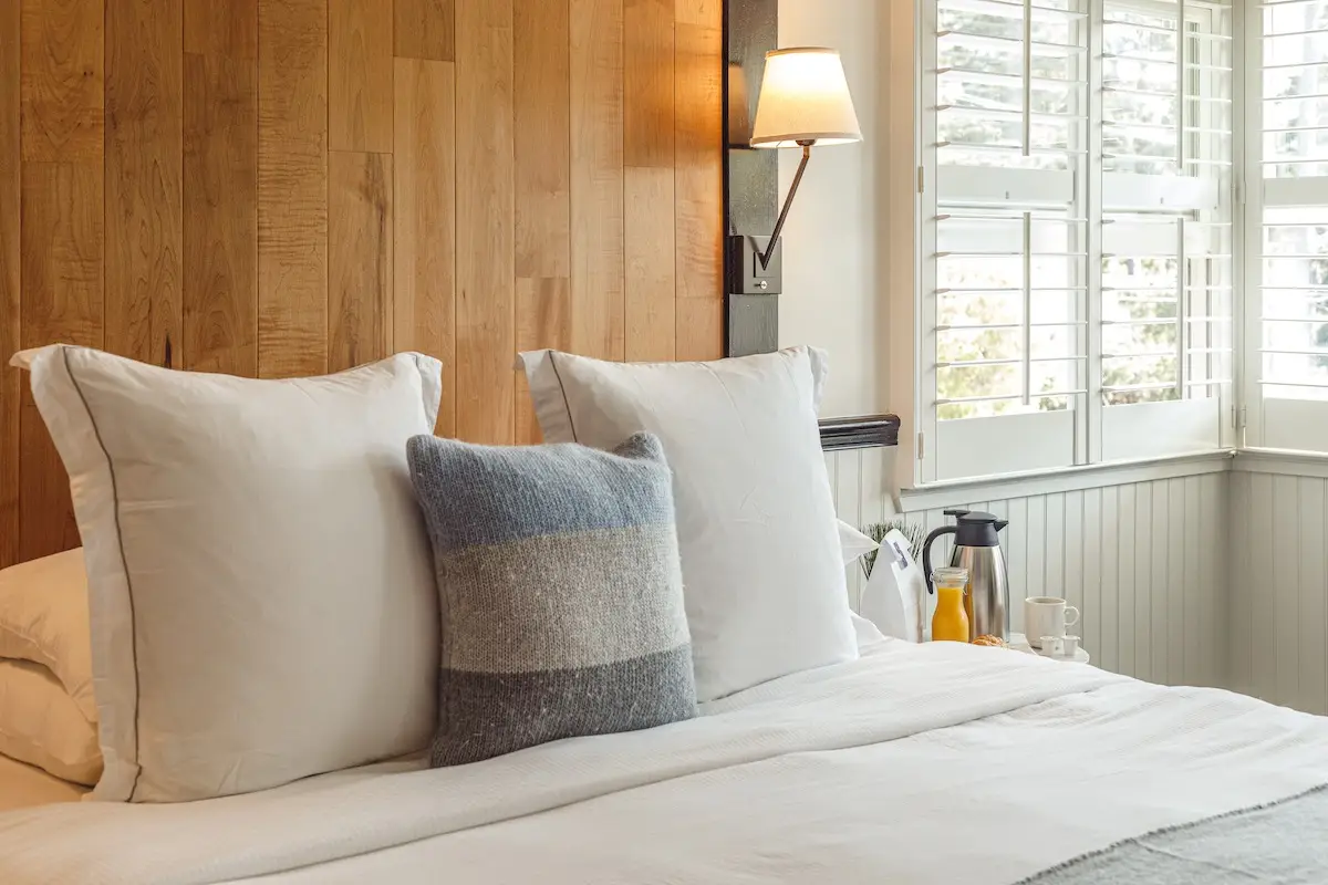 Cozy bedroom with a wooden accent wall, two white pillows, and a gray-blue striped cushion on a neatly made bed. A bedside wall lamp illuminates the space, and a window with white blinds allows natural light to filter in. A coffee set is placed by the window, making this room feel like The Hideaway.