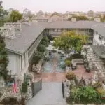Aerial view of The Hideaway's courtyard within an L-shaped building surrounded by greenery. The charming Carmel courtyard features outdoor seating areas with umbrellas, stone pathways, and a U.S. flag near the entrance gate. Trees and shrubs are present throughout the premises.